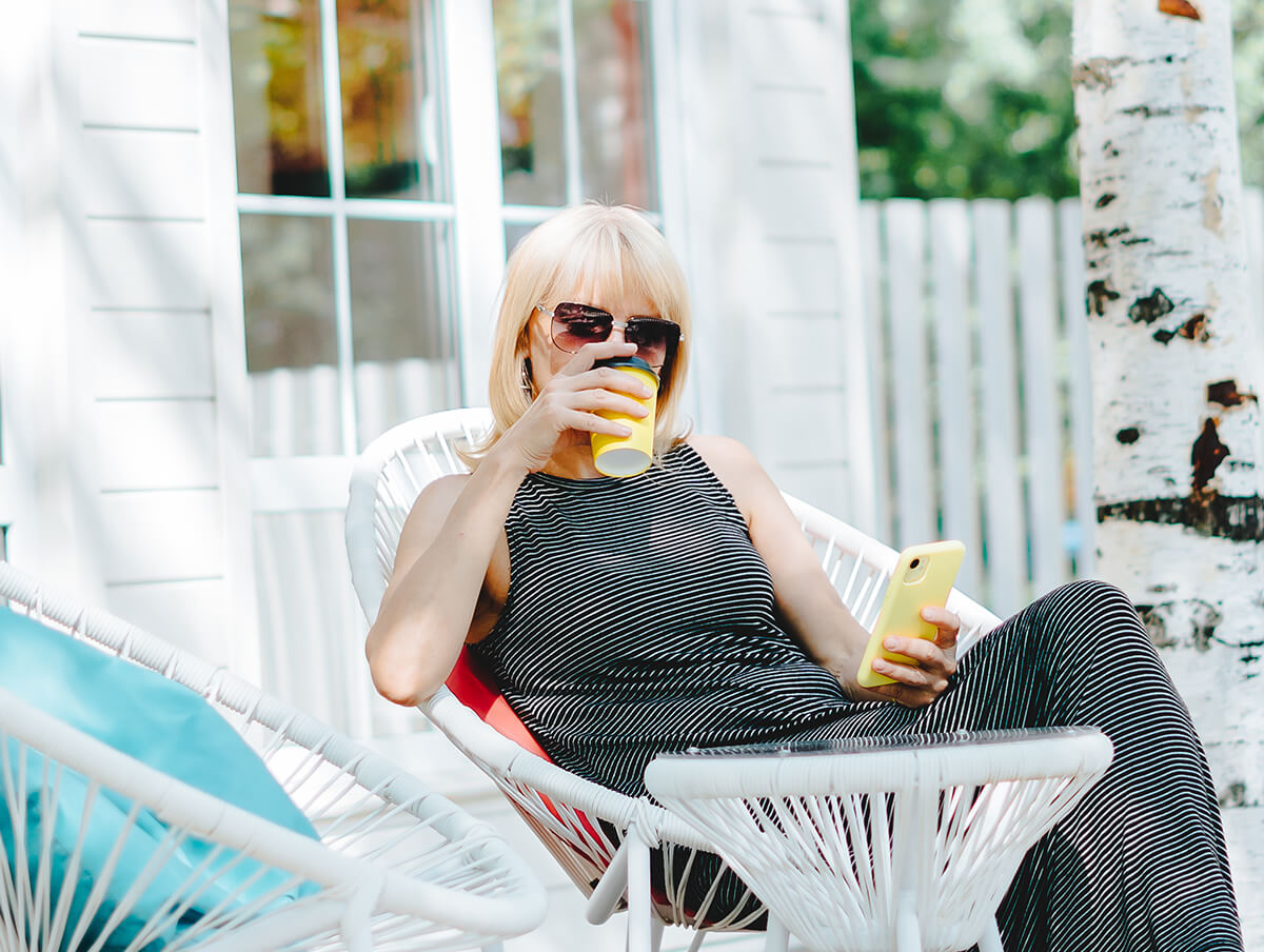 A woman in a black dress drinking coffee while looking at her yellow cell phone.